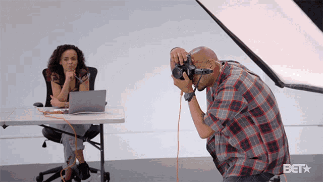 a man in a plaid shirt is taking a picture of a woman sitting at a table with a laptop on it