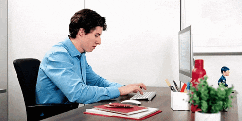 a man in a blue shirt sits at a desk typing on a computer