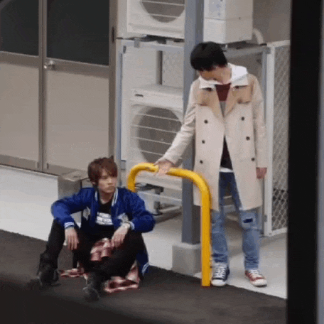 a man in a trench coat is standing next to another man sitting on the ground .