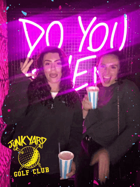 two women holding drinks in front of a neon sign that says do you