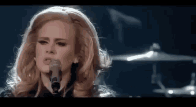 a woman is singing into a microphone with a drum set in the background