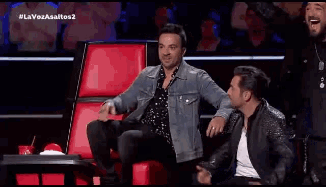 a man in a denim jacket is sitting in a red chair with other men .