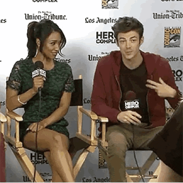 a man and a woman are sitting in chairs talking into microphones . the man is wearing a shirt that says hero .