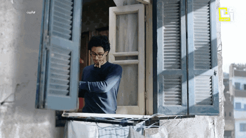 a man in glasses looks out of a window with shutters and a foreign language written on the bottom