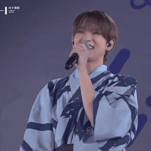 a man wearing a blue and white kimono is singing into a microphone with the hybe label on the bottom