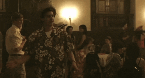 a man wearing a hawaiian shirt and a feathered hat stands in a crowded room
