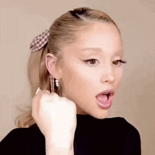 ariana grande is wearing a ponytail and earrings and is making a fist with her tongue out .
