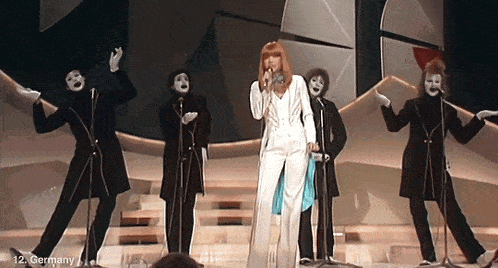 a woman singing into a microphone in front of a group of mimes with germany written on the bottom right