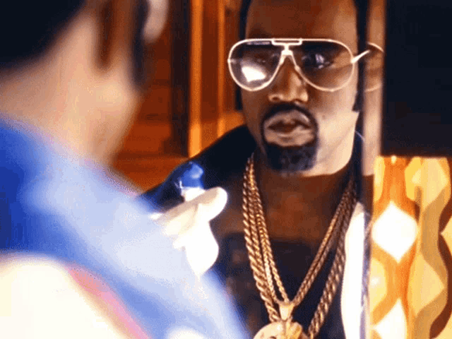 a man wearing sunglasses and a gold chain looks at himself in a mirror