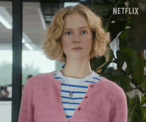 a woman wearing a pink sweater and a striped shirt is standing in front of a plant .