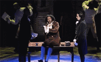 a man in a costume is sitting at a table in a dark room while two other men stand around him .