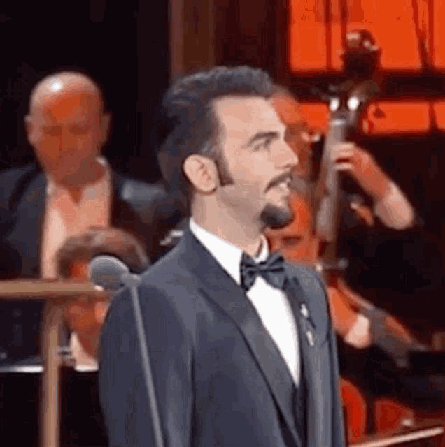 a man in a suit and bow tie is standing in front of a microphone in front of an orchestra .