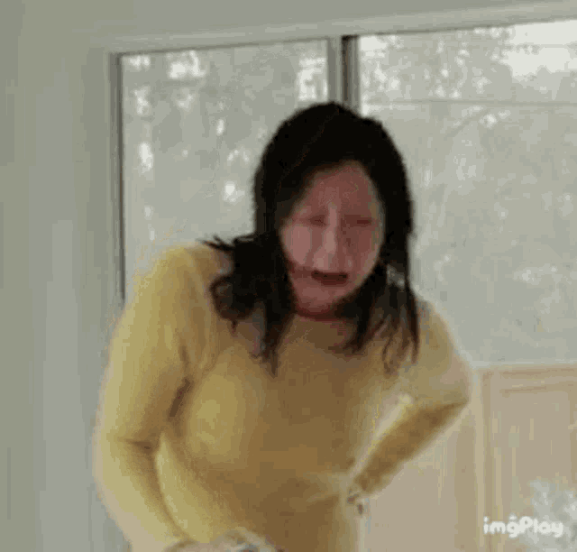 a woman in a yellow sweater is standing in front of a window with her eyes closed .