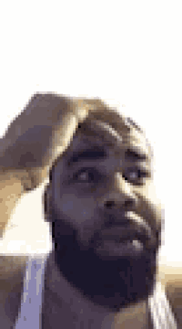 a man with a beard is wearing a white tank top and scratching his head .