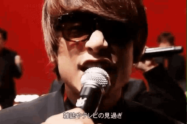 a man wearing sunglasses is singing into a microphone while another man holds a gun behind him .