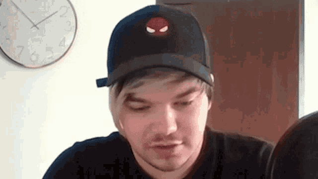 a man wearing a black hat and a black shirt is sitting in front of a wall clock .