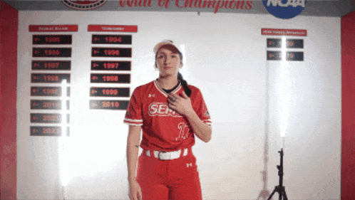 a woman in a softball uniform stands in front of a wall that says wall of champions