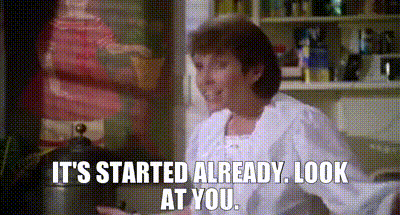a woman is sitting in a kitchen with the words `` it 's started already look at you '' written on the screen .