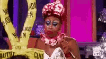 a drag queen is wearing a crown and holding a microphone while standing in front of a pink wall .