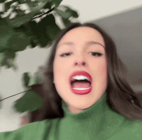 a woman wearing a green turtleneck is making a funny face with her mouth open
