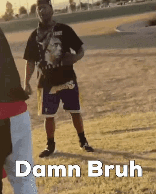a man in a black shirt and purple shorts is standing in a field with the words damn bruh written below him .