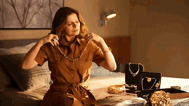 a woman in a brown shirt is sitting on a bed looking at her jewelry