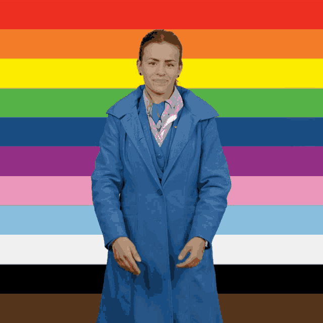 a woman wearing a blue coat stands in front of a rainbow flag