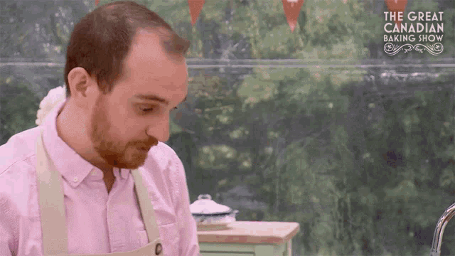 a man in a pink shirt and apron is standing in front of a sign that says the great canadian baking show