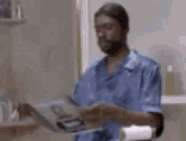 a man in a blue shirt is standing in a bathroom holding a book .