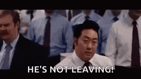 a man in a suit and tie is standing in front of a crowd of people and says `` he 's not leaving ! ''
