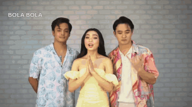 a woman in a yellow dress stands between two men in front of a brick wall that says bola bola on it