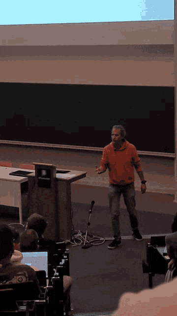 a man in an orange shirt is giving a presentation in front of a projector screen