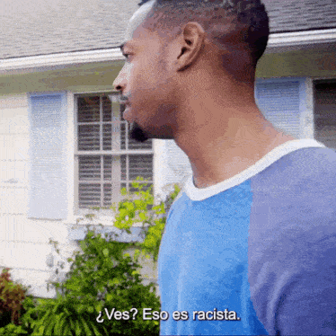 a man in a blue shirt stands in front of a house and says " ves "