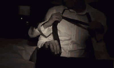 a man in a white shirt and black tie is sitting on a bed in a dark room .