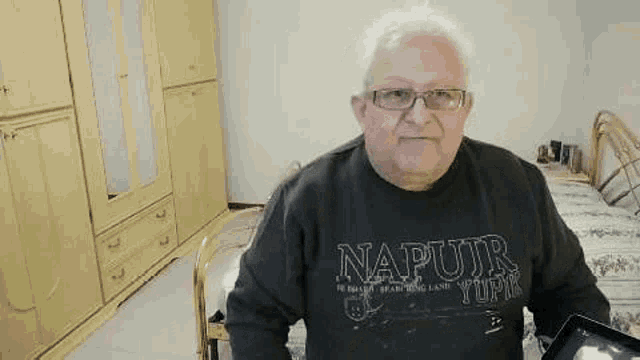 an elderly man wearing glasses and a napurr shirt is sitting in a bedroom holding a tablet computer .