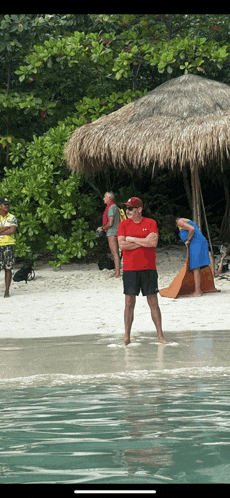a man in a red shirt is standing on a beach with his arms crossed