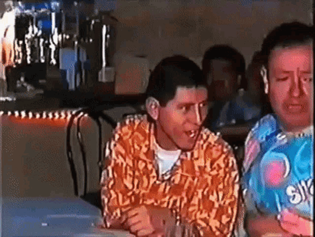 two men are sitting at a table with one wearing a shirt with the letter s on it .