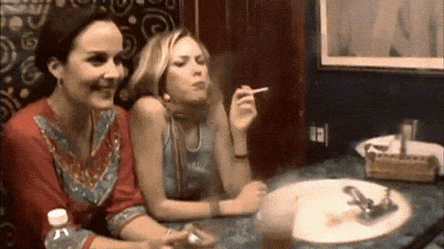 two women are sitting at a table one is smoking a cigarette and the other is drinking a drink