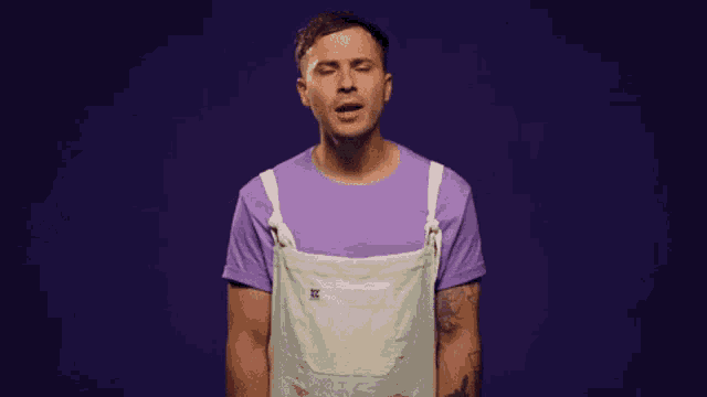 a man wearing a purple shirt and white overalls is making a funny face .