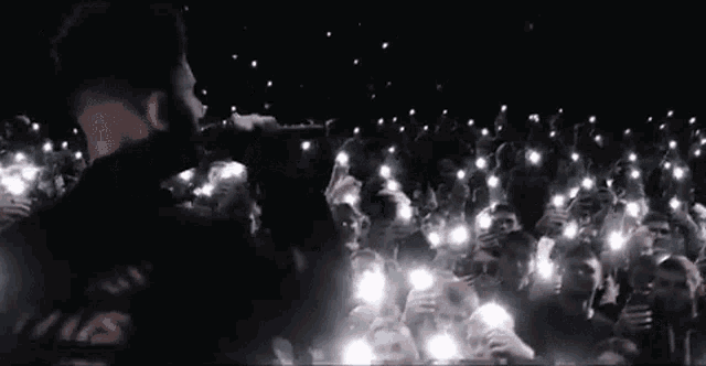 a man is singing into a microphone in front of a crowd of people holding their cell phones .