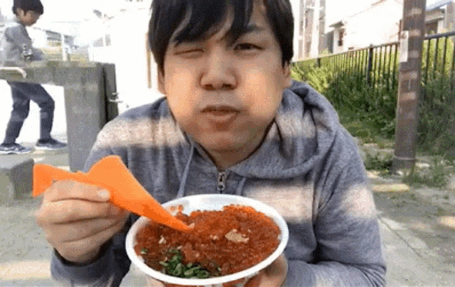a man in a grey hoodie is eating a bowl of food with an orange spoon .