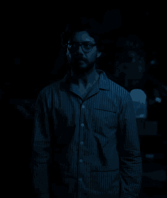 a man in a blue striped shirt and glasses stands in the dark