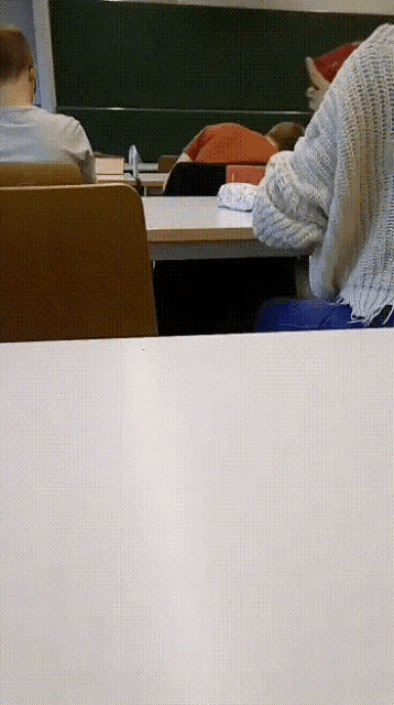 a group of people sit at desks in a classroom with a green board behind them