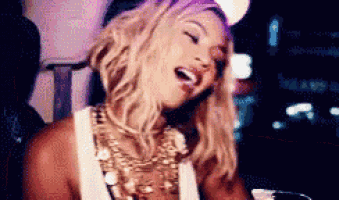 a woman wearing a white tank top and gold jewelry is laughing while standing in a dark room .