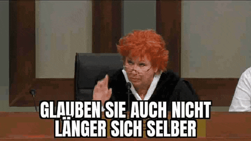 a woman with red hair is sitting at a table with a microphone and a sign that says glauben sie auch nicht