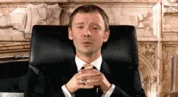 a man in a suit and tie sits in a chair with his hands folded