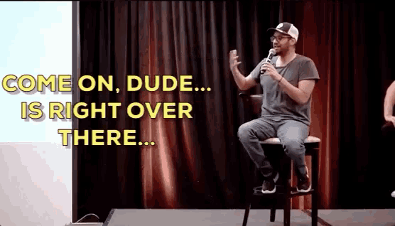 a man sitting on a stool holding a microphone with the words come on dude is right over there