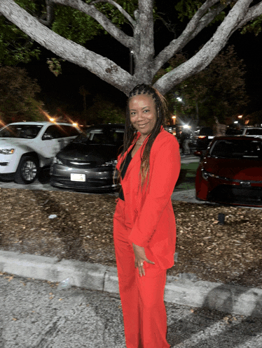 a woman in a red suit is standing in front of a tree and cars