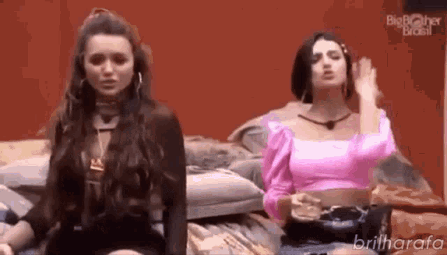 two women are sitting next to each other on a bed . one of the women is wearing a pink top .