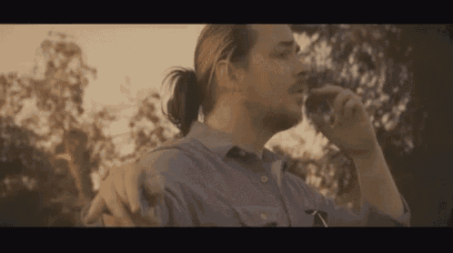 a man with a ponytail and a mustache is smoking a cigarette in a park .